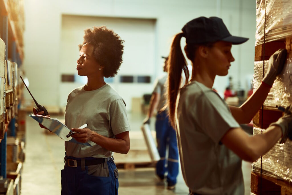 Black female inspector with walkie-talkie checking stock of products while working in a warehouse.